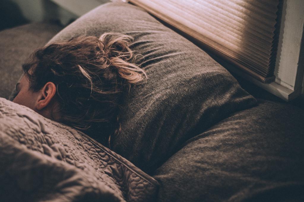 A woman is asleep and part of her face and brown hair is visible - her head is on a brown pillow and she os covered by a brown blanket. Part of a window covered by a brown blind is visible behind the pillow. 